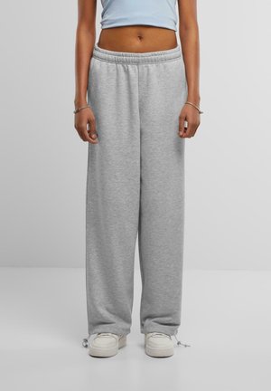 Persona vistiendo pantalones deportivos sueltos de color gris claro y zapatillas blancas, de pie frente a un fondo gris liso.
