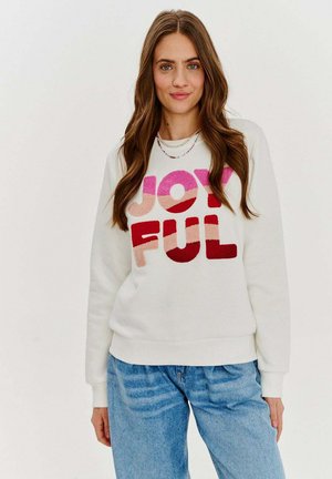 Bílý sweatshirt s kulatým výstřihem s nápisem "JOYFUL" v mnoha barvách a texturovaném písmu. Kombinovaný s světle modrými džíny.