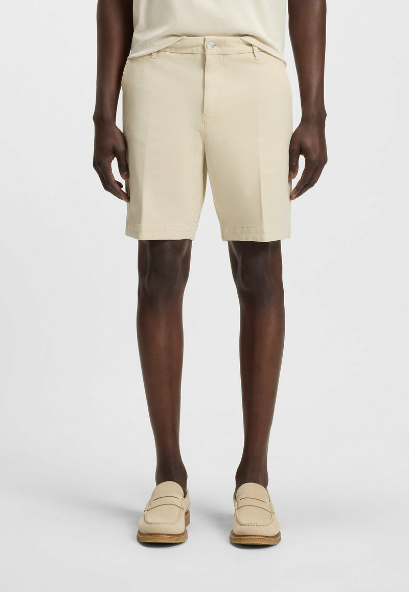 Pantaloni corti beige in cotone con davanti piatto, due tasche laterali e una texture liscia, abbinati a mocassini chiari. Design semplice e classico.