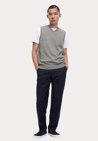 Maglione senza maniche grigio sopra una maglietta bianca, abbinato a pantaloni scuri a righe sottili e sneakers blu navy. Presentazione di un outfit semplice e informale.