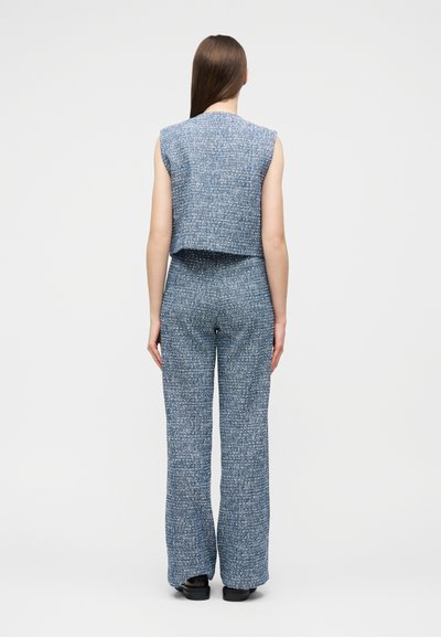 Γυναίκα με μακριά καστανά μαλλιά, που φοράει μπλε αμάνικη μπλούζα με υφή και ταιριαστό παντελόνι φαρδιάς γραμμής, στέκεται με την πλάτη στραμμένη σε απλό φόντο.