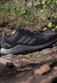 Chaussure de trail Adidas Terrex gris foncé sur un sol forestier rocheux avec des feuilles de plantes vertes en arrière-plan.