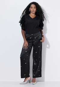 Černá blůza s vrstvenými krajkovými rukávy, v kombinaci s lesklými černými kalhotami širokého střihu se stříbrnými ozdobami, a stříbrnými podpatky.