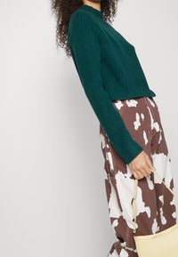 Vrouw met een donkergroen geribbeld trui en bruin-witte gebloemde broek, die een kleine lichtgele handtas aan het hengsel vasthoudt.