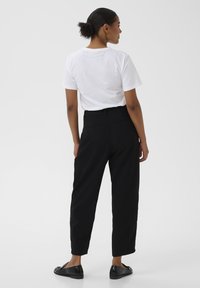 Femme avec un chignon bouclé portant un T-shirt blanc à manches courtes, un pantalon noir taille haute fuselé et des mocassins noirs, se tenant de dos sur un fond blanc.