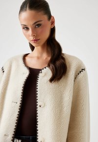 Giacca in shearling bianca con scollatura tonda, bordature ondulate marroni e bottoni frontali, indossata sopra un top marrone. Texture liscia, design casual.