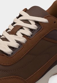 Καφέ αθλητικό παπούτσι με λευκά κορδόνια, πάνελ από σουέντ και ύφασμα, και ραμμένες λεπτομέρειες κατά μήκος της πλευράς και γύρω από τις τρύπες των κορδονιών.