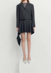 Dark gray cropped blazer over a matching flared skirt. Model holds a black bag, wearing white pointed-toe heels. Smooth texture.
