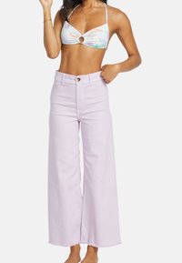 Top de bikini con un estampado colorido de hojas de palma y un corte circular, combinado con pantalones de pierna ancha de cintura alta en color lavanda con dobladillos sin rematar.