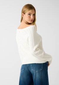 Pull en maille torsadée blanche avec un design épaules dénudées, ourlet côtelé et coupe décontractée, associé à un jean bleu à jambes larges.