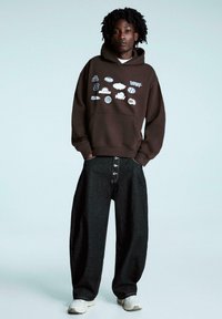 Brązowa bluza z kapturem z różnorodnymi graficznymi logo, zestawiona z luźnymi, ciemnymi dżinsowymi spodniami z dużymi guzikami i białym przeszyciem.