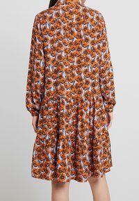 Robe fleurie à manches longues, avec des fleurs orange sur un fond bleu clair. Présente un design à volants et des poignets froncés. Texture de tissu douce.