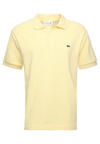 Žluté polo tričko z texturované bavlny, s pečlivě složeným límečkem a malým zeleným logem Lacoste na levé straně hrudi.