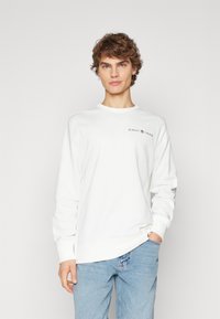 Hvid bomulds sweatshirt med rund halsudskæring, lange ærmer og et lille sort broderet logo på brystet. Båret sammen med lyseblå jeans.