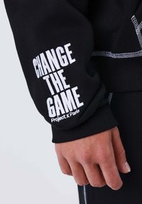 Sudadera negra con texto bordado en blanco "CAMBIA EL JUEGO" y "Project x Paris" en la manga; presenta un puño texturizado.