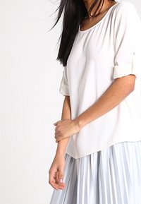 White short-sleeve blouse with a rounded neckline and rolled sleeves, paired with a pleated light blue skirt. Smooth fabric texture.