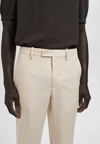 Beige Tailored-Hosen mit einer glatten Textur, ausgestattet mit einem Gürtelschlaufen und einem Knopfverschluss. Getragen mit einem dunkelbraunen, kurzärmeligen Hemd.