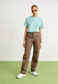 T-shirt en coton bleu clair avec un petit graphisme, associé à un pantalon taille haute marron. Baskets blanches, cheveux texturés et lunettes de soleil.