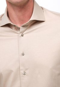 Beige Hemd mit Knopfleiste, weicher Textur, tailliertem Schnitt und spitzen Kragen. Verfügt über silberne Knöpfe entlang der Vorderseite.