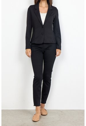 Femme portant un blazer noir cintré, un jean slim noir, un haut blanc orné de dentelle, et des mocassins beiges, debout devant un fond blanc.