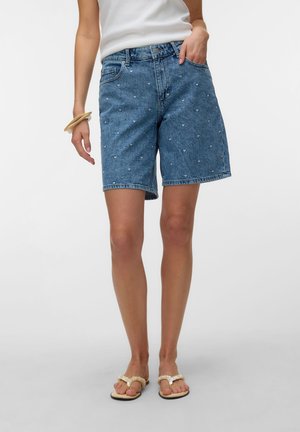 Femme portant un short en jean bleu avec de petits motifs en forme de cœur, un haut blanc sans manches, des sandales beige et un bracelet beige au poignet gauche.