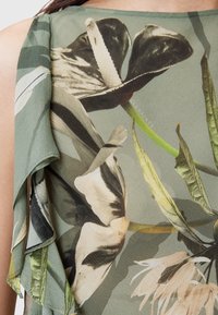 Gros plan sur un tissu vert avec un grand imprimé floral dans des teintes de noir, crème et vert, présentant un détail de volants superposés sur une épaule.