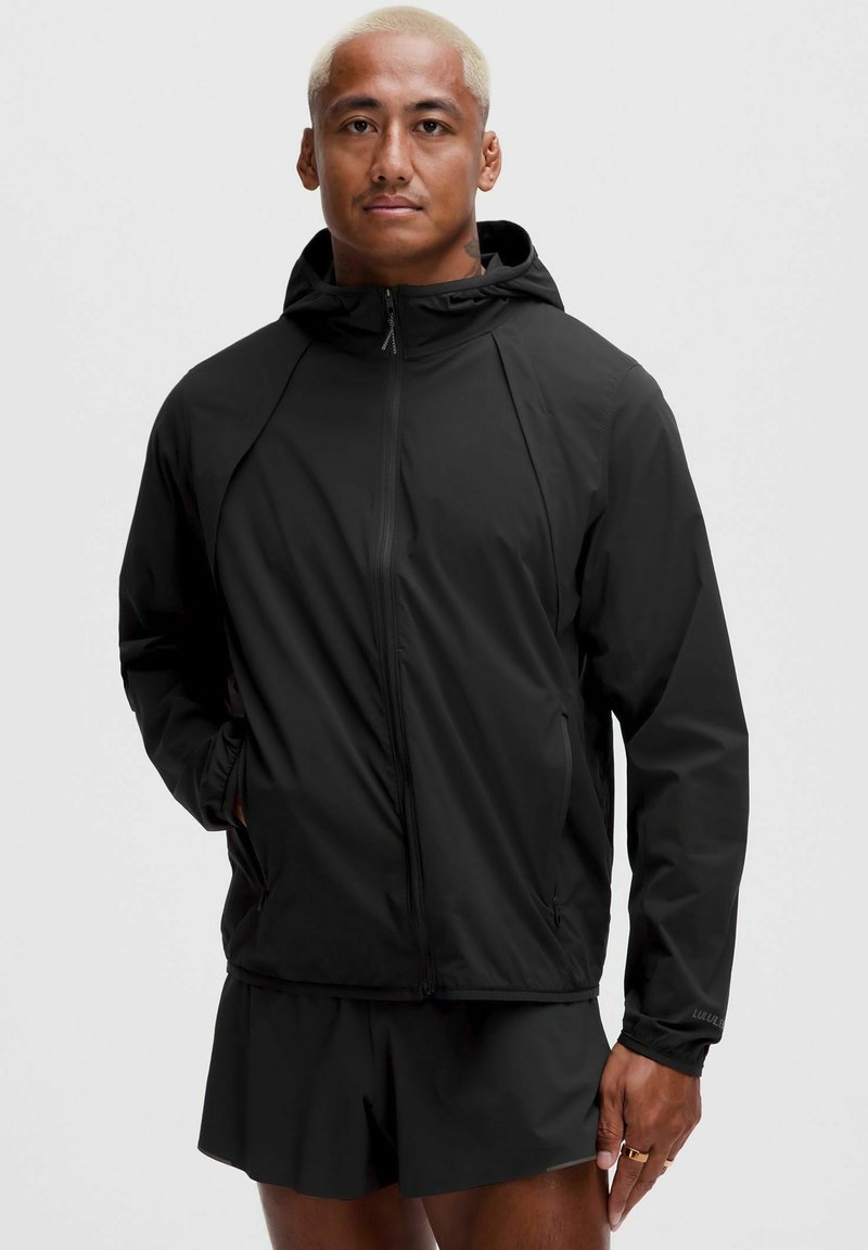 Vīrietis ar īsiem blondiem matiem, valkājot melnu kapuci un melnas šortus, stāv ar vienu roku kabatā vienkāršas aizmugures priekšā.