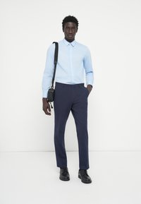 Svetlo modra srajca z dolgimi rokavi, v kombinaciji s temno mornarsko hlačami in črnimi usnjenimi čevlji. Črna torba visi z ramena.