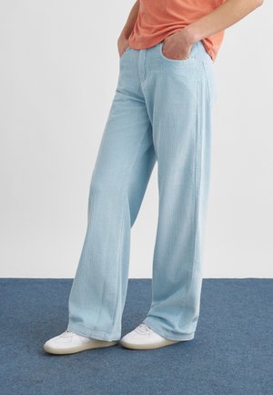 Osoba ubrana w jasnoniebieskie szerokie spodnie sztruksowe, białe trampki i pomarańczową koszulę, stojąca na niebieskim dywanie z rękami w kieszeniach.