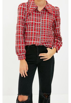 Persoon die een rode geruite blouse met lange mouwen, opgepofte schouders en zwarte gescheurde spijkerbroek draagt, staand tegen een witte achtergrond.