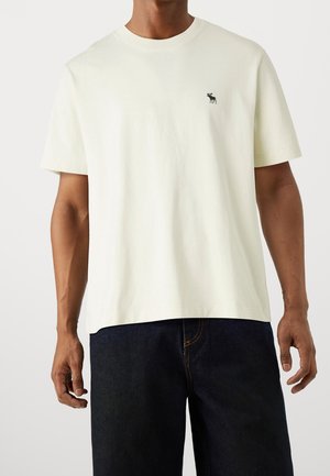 Homme portant un t-shirt à manches courtes uni de couleur blanc cassé avec un petit logo d'orignal foncé sur la poitrine gauche, associé à un jean bleu foncé.