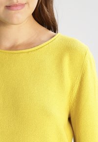 Gelbes Strickpullover mit Rundhalsausschnitt und strukturiertem Stoff, der ein geripptes Muster und mittlere Dicke zeigt.