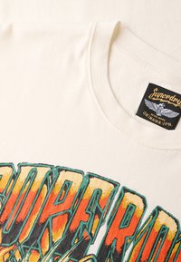 Camiseta de algodón color crema con texto gráfico vibrante en tonos de naranja, amarillo y azul verdoso, que presenta una etiqueta negra con la marca en el cuello.