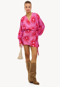 Vestido cruzado rosa con patrones de rayos de sol rojos, mangas largas y cintura anudada. Combinado con botas altas de ante marrón y un bolso crema.