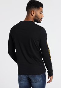 Black long-sleeve shirt with a round neck, featuring a yellow graphic design on the left sleeve. Made of smooth fabric, fitted silhouette.