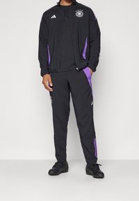 Zwarte sportieve trainingspak met paarse accenten. Het zip-up jack heeft logo's en contrasterende strepen; bijpassende broek met elastische tailleband.