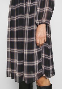 Robe à manches longues à carreaux noirs, beiges et bleus avec poignets froncés, portée avec des bottes noires montantes jusqu'aux genoux, montrée de la mi-cuisse à la main.