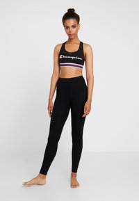 Sujetador deportivo negro con logo blanco de 'Champion' y acentos en rojo y azul, combinado con leggings negros de cintura alta; tejido suave, diseño ajustado.