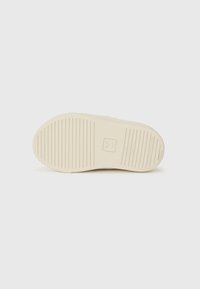 Sål av sko med en teksturert, ribbet design i lys beige, med synlig logoavtrykk og flat profil for stabilitet.