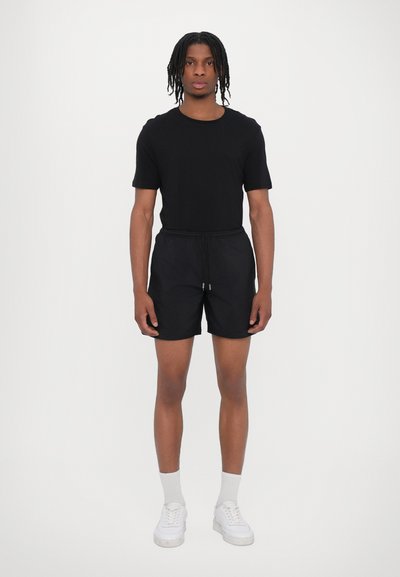 Schwarzes T-Shirt mit kurzen Ärmeln, kombiniert mit schwarzen Shorts mit elastischem Bund. Getragen mit weißen Sneakers und Socken in Knöchellänge.