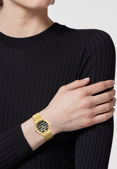 Kuldse metallist käekell mustade triipudega sihverplaadiga, ümarate numbritega ja soonikuga kuldse käevõruga, kantud palja randme peal musta tekstuurse kootud kampsuni vastu.