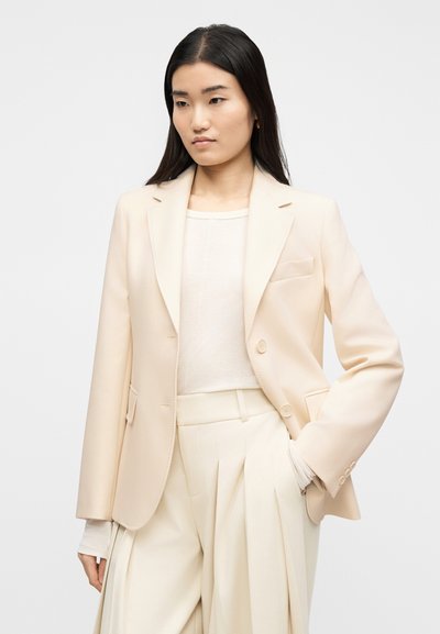 Γυναίκα με μακριά μαύρα μαλλιά φοράει κρεμ σακάκι, λευκή μακρυμάνικη μπλούζα και κρεμ ψηλόμεση πλισέ παντελόνα, με το ένα χέρι στην τσέπη.