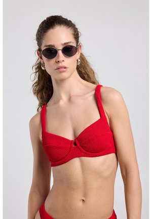 Frau mit langen lockigen Haaren, die silberne Creolen, schwarze runde Sonnenbrillen und ein strukturiertes rotes Bikinioberteil vor einem einfarbigen hellen Hintergrund trägt.