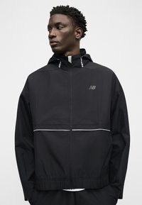 Άνδρας που φοράει μαύρο μπουφάν New Balance με φερμουάρ, κουκούλα, ανακλαστικές ρίγες και ελαστική μέση, κοιτάζοντας αριστερά σε απλό φόντο.