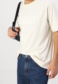 Sandfärgad bomull T-shirt med präglad "COACH"-logotyp, i kombination med mörkblå denimjeans. Slim-fit design, avslappnad stil. Svart axelrem över axeln.