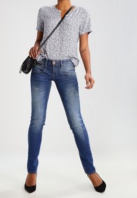 Kortärmad mönstrad blus i vitt och marinblått som är kombinerad med figurnära blåjeans. Accessoarer inkluderar en liten svart crossbody-väska.