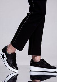 Osoba ubrana w czarne spodnie z srebrnym paskiem oraz czarne trampki z białymi podeszwami i czarną gwiazdą z boku.