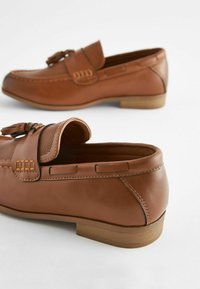 Mocassins en cuir marron avec un dessus texturé, des franges décoratives et une semelle plate couleur marron clair. Les coutures mettent en valeur les détails du design.