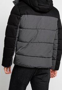 Piumino con schema di colori nero e grigio, design trapuntato e cappuccio. Realizzato in materiale leggero con una texture opaca.