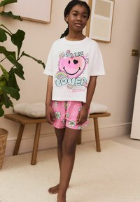 T-shirt blanche avec un graphique de visage souriant rose et un texte coloré, associée à un short rose avec un motif floral comportant des visages souriants.